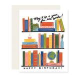 A delightful birthday card featuring beautifully illustrated bookshelves adorned with colorful books. Among the shelves, a special touch includes a birthday cake, a globe, and a house plant – a perfect celebration for the avid book lover. The card reads, 'May it be a year for the books. Happy Birthday! 