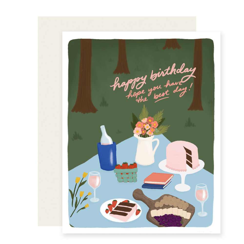 A delightful birthday card with charming illustrations of a scenic picnic surrounded by a lush forest. The picnic blanket is set with flowers, a bottle of wine, a birthday cake, and a tempting charcuterie board – a perfect setting for birthday celebrations amidst nature's beauty.The card reads 'Happy Birthday. Hope you have the best day!