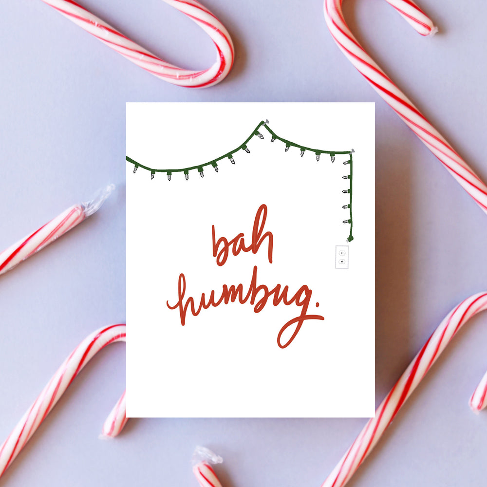  A cheeky holiday card with a minimalist design, featuring a single strand of twinkling lights draped across the top. In bold red letters, the card proclaims 'Bah Humbug,' capturing a playful, festive spirit for those with a humorous take on the season.