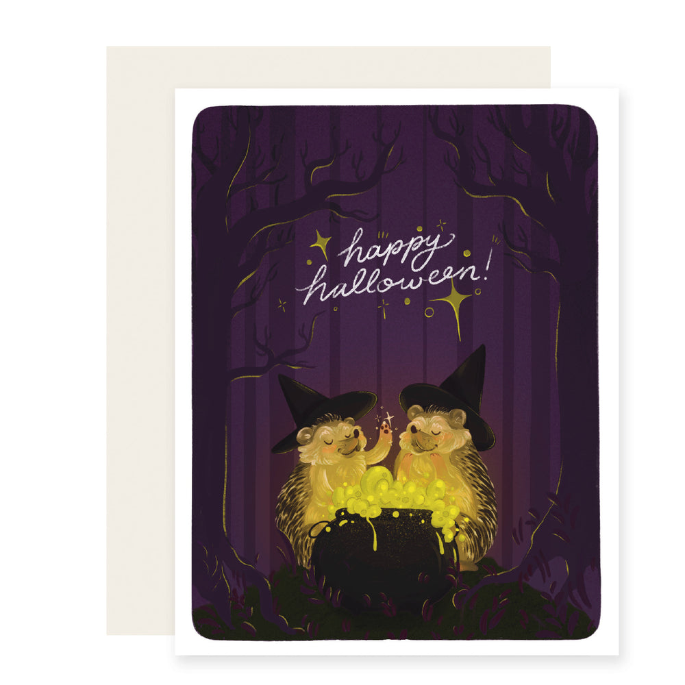 A charming Halloween card featuring two hedgehogs wearing witches' hats, brewing a potion. The card is colorfully illustrated and reads 'Happy Halloween.'