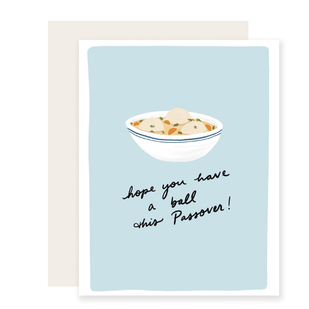 A Passover card with a light blue background featuring an artfully illustrated bowl of matzo ball soup. The card reads, 'Hope you have a ball this Passover,' creating the perfect blend of puns, warmth, and smiles. 