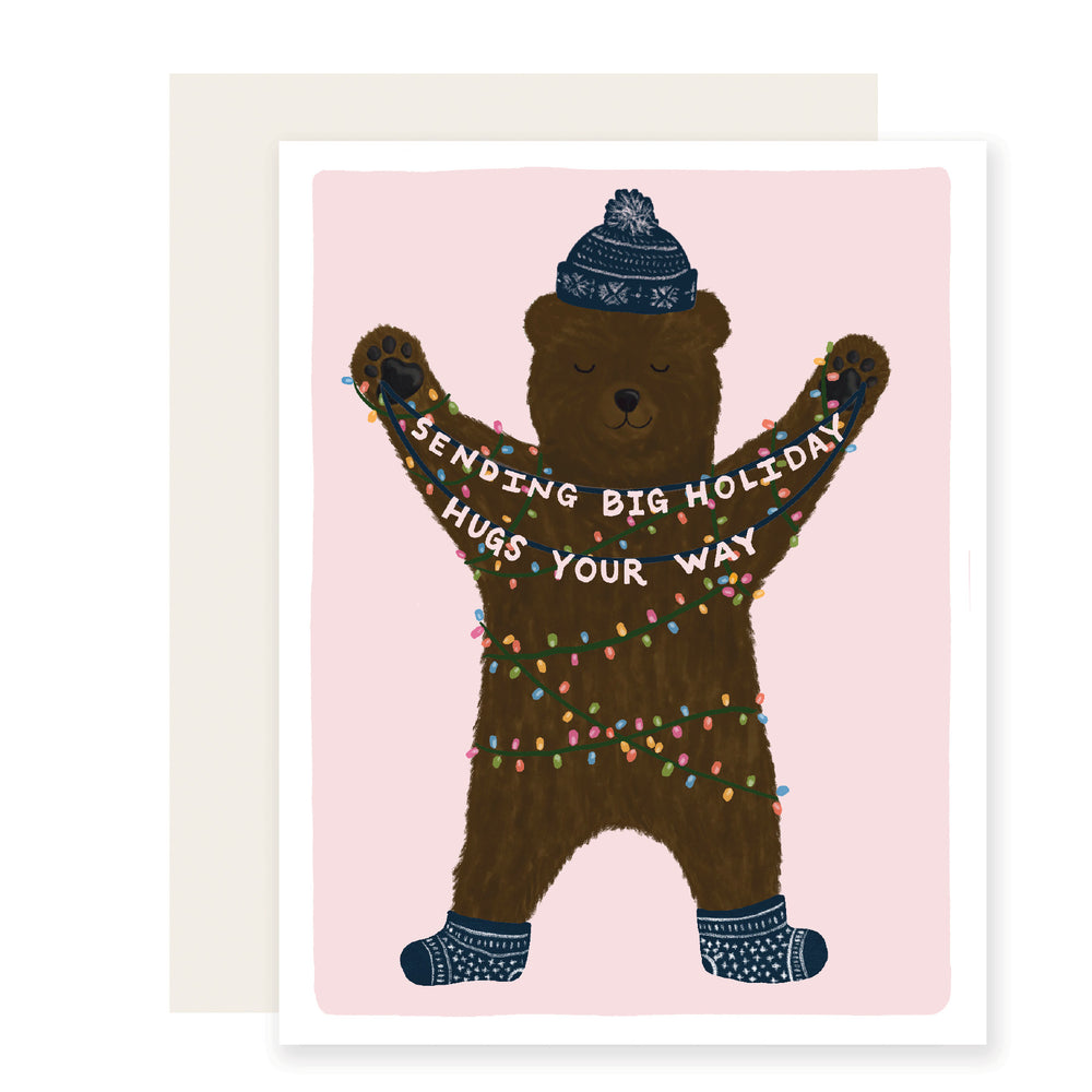 An endearing holiday card with an adorably  illustrated bear wrapped in holiday lights and wearing a cozy knit winter hat and socks. The bear holds a banner that reads Sending big holiday hugs your way adding a heartwarming touch to the festive scene